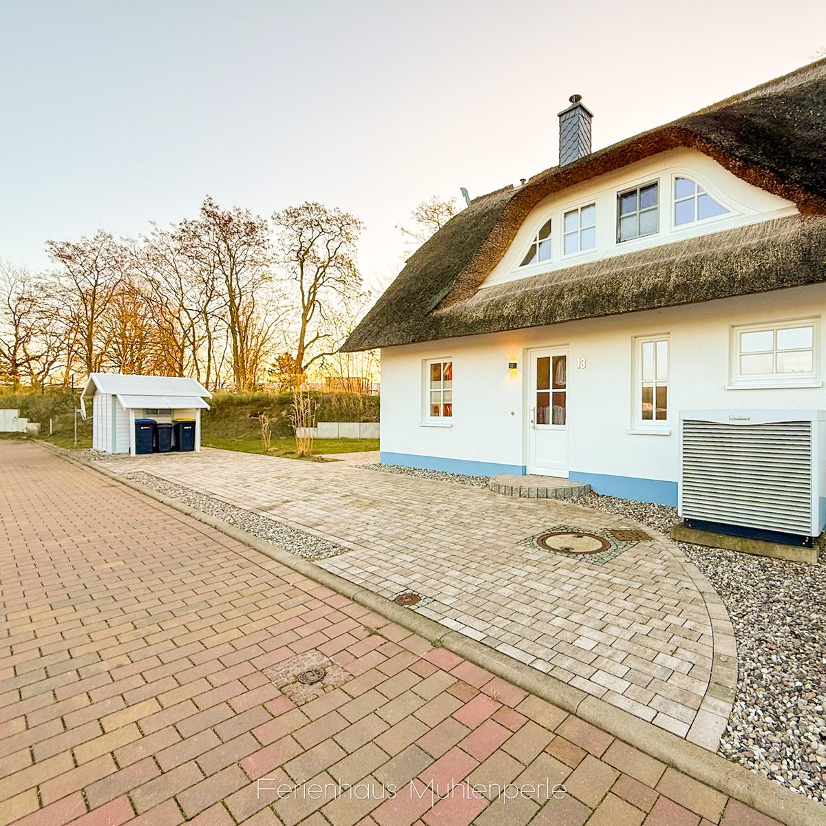 Reetgedecktes Ferienhaus Mühlenperle mit gepflastertem Eingangsbereich bei Sonnenuntergang.